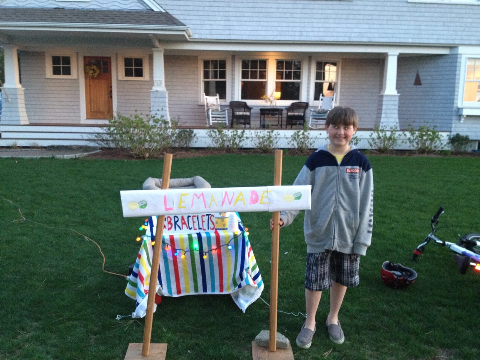 Joey at nine beside a hand-painted “Lemanade” sign and a striped table of bracelets strung with colored lights, on a lawn in front of a gray-shingled porch.