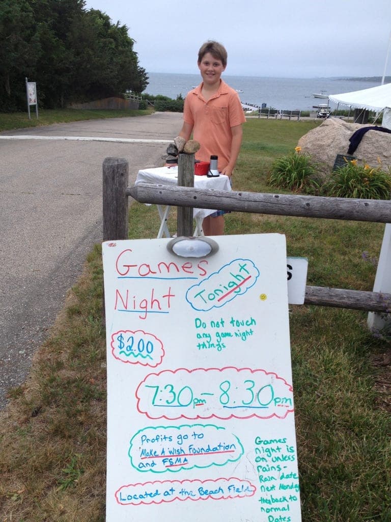 Joey behind a small table by the water, with a hand-lettered whiteboard: Games Night tonight, $2, 7:30–8:30 pm at the Beach Field, profits to the Make-A-Wish Foundation.