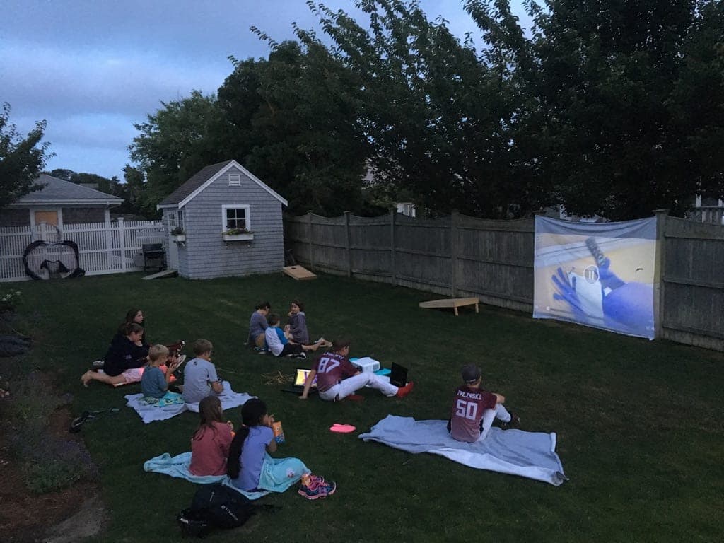 Kids on blankets in a backyard at dusk, watching a movie projected onto a sheet hung on a wooden fence.