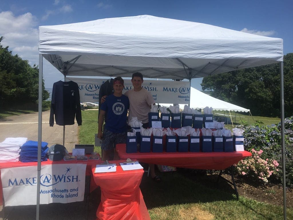Joey and a friend under a white tent with Make-A-Wish banners, behind tables of folded T-shirts, a hung sweatshirt and rows of navy gift bags.