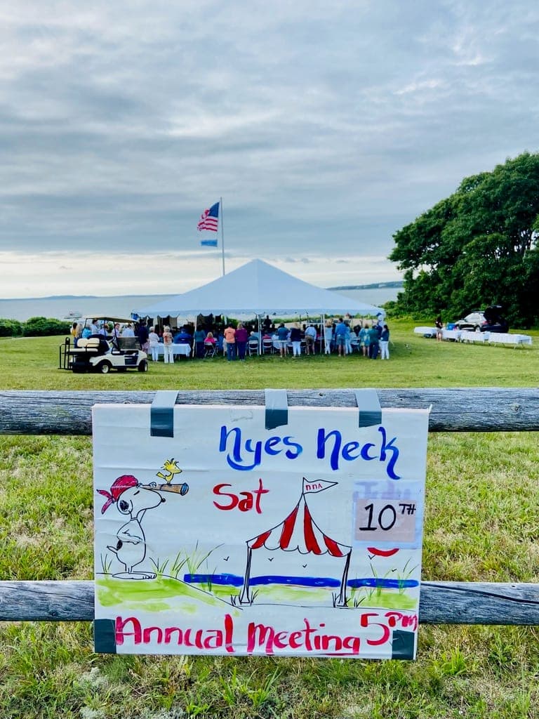 A hand-drawn sign on a split-rail fence reading “Nyes Neck, Sat 10th, Annual Meeting 5pm”, with a crowd gathered under a white tent by the water behind it.