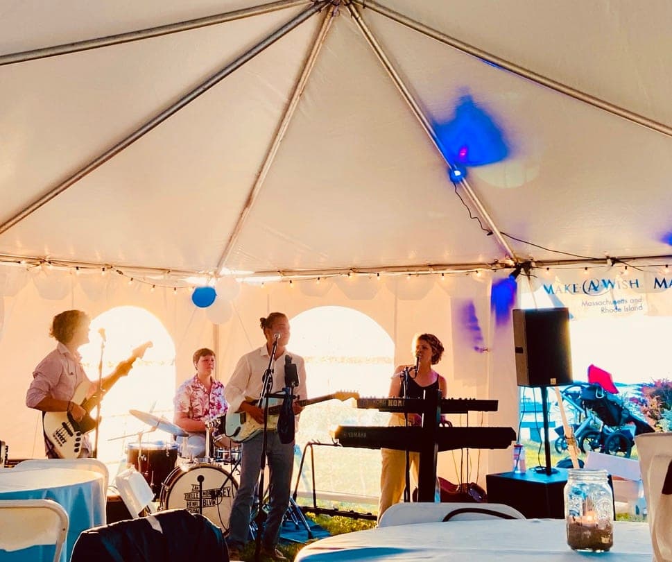 A four-piece band — bass, drums, guitar and keyboard — playing under a white tent strung with lights and a Make-A-Wish banner.