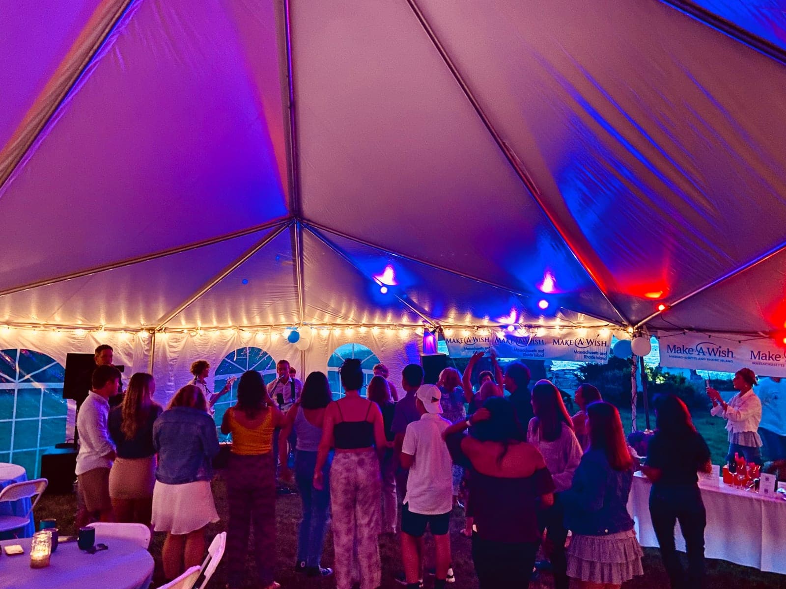 A crowd dancing under a tent lit purple and blue at night, with Make-A-Wish banners along the back wall.
