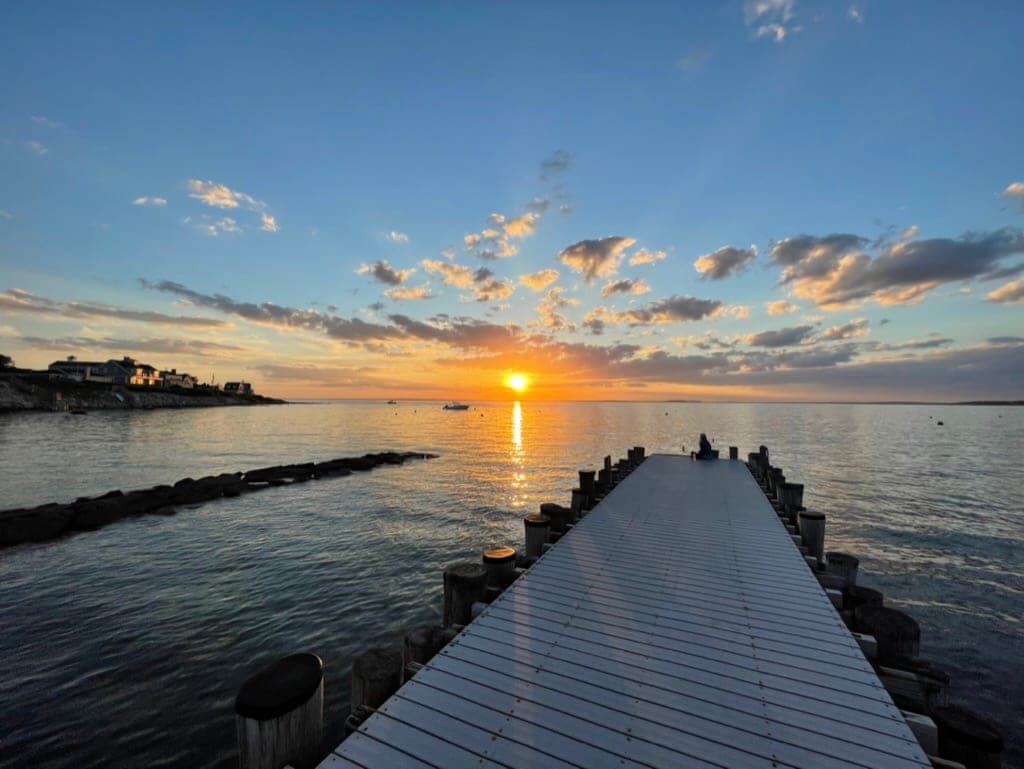 The sun setting over calm water at the end of a long wooden dock, one person sitting alone at the far end, a jetty of rocks and shingled houses on the point to the left.
