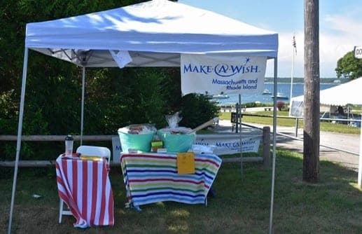 A white pop-up tent with a Make-A-Wish Massachusetts and Rhode Island banner over two striped tables, with the harbor behind it.